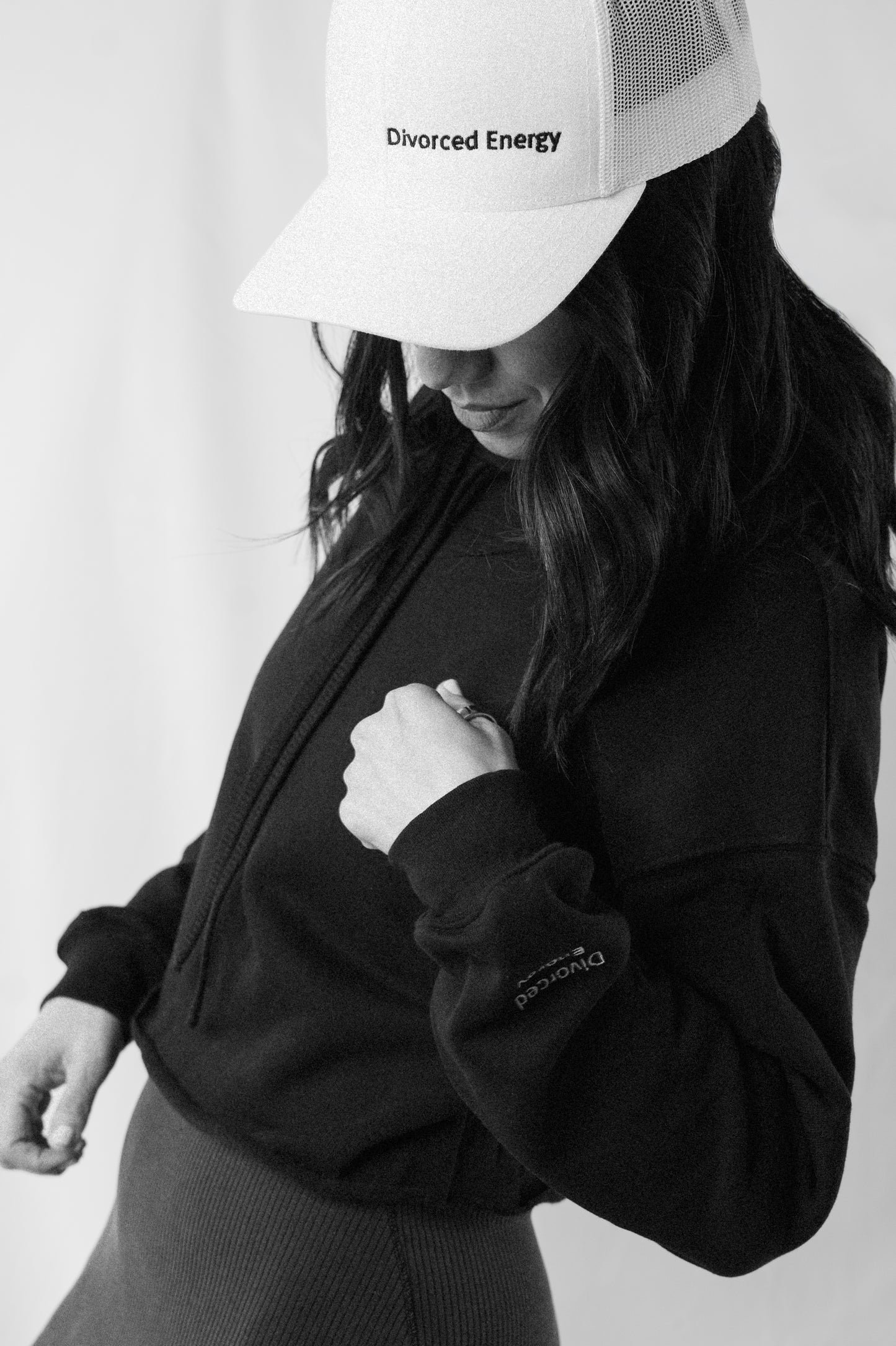 Person wearing a black hoodie and white cap with text, standing against a plain background